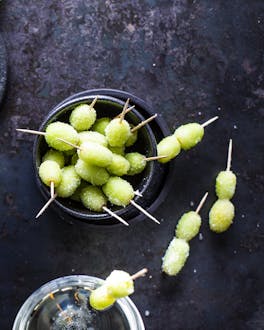 Gefrorene grüne Trauben an Zahnstochern stehen in Schälchen vor dunklem Hintergrund neben einer Flasche Prosecco und einem Glas Wodka.