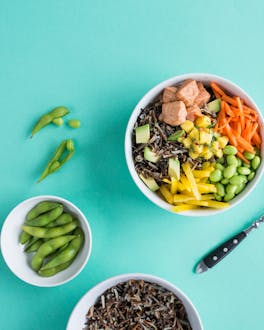 Bowl mit schwarzem Reis, Mangochutney, Avocado, Edamame, Karottenstreifen und Lachsstücken auf türkisem Untergrund