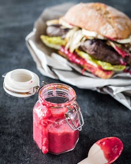 Pinkfarbenes Beeren Ketchup in einem Weckglas mit Deckel neben einem Burger und einem Holzlöffel auf grauem Untergrund.