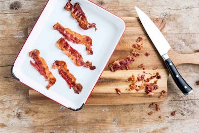 5 Stücke Bacon liegen auf einem Teller auf einem Holztisch, daneben ein Messer und kleine Stücke Bacon