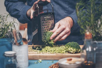 Hände verarbeiten Brokkoli-Röschen mit einer Küchenreibe auf Holzbrett