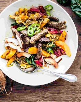 Rinder-Curry mit buntem Gemüse auf einem weißen Teller auf einem Holztisch, daneben als Deko eine halbe Kokosnuss.
