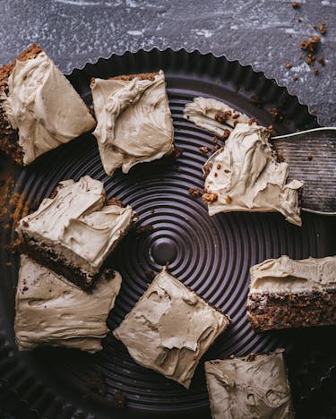 Stücke von Carob-Brownies auf einer dunklen Servierplatte, ein Stück liegt auf einer Kuchenschaufel. Fotografiert von oben auf dunklem Untergrund, etwas Carob-Pulver ist noch verstreut.