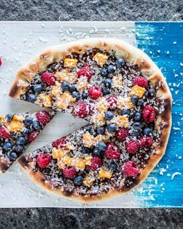 Angeschnittene Pizza von oben mit Schokolade bestrichen und mit Früchten belegt auf blau-weißem Holzbrett und grauem Untergrund.