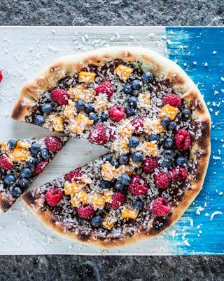 Angeschnittene Pizza von oben mit Schokolade bestrichen und mit Früchten belegt auf blau-weißem Holzbrett und grauem Untergrund.