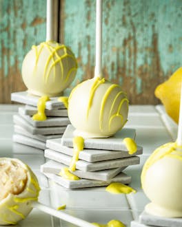 Aufkleinen Türmchen aus Unterlegern sind hellgelbe Cake-Pops drapiert. Drum herum ist ein wenig gelbe Glasur verteilt.