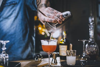 Eine tättoowierte Hand gießt den Trinidad Sour durch einen Strainer in eine Cocktailschale.