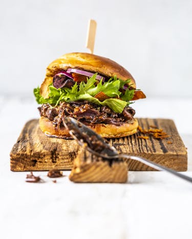 Auf einem Holzbrett vor weißem Hintergrund liegt in Burger mit Jackfruit, Salat und Zwiebelringen. Davor lehnt ein Esslöffel.