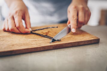Eine Vanilleschote wird mit einem Messer auf einem Holzbrett ausgeschabt.