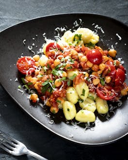 Gnocchi mit Tomatensauce und Kichererbsen auf dunklem Teller, daneben ein Schälchen Parmesan