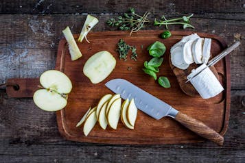 Auf einem Holzbrett liegt ein Stück Apfel, Apfelscheiben, ein großes Küchenmesser, Basilikum und eine Rolle Ziegenkäse.