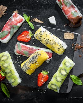 In fruchtigen Farben sind die mit Kiwi, Erdbeeren und Mango gefüllten Sommerrollen auf einem schwarzen Brett verteilt. Dabei steht eine Schale mit Schokosauce.