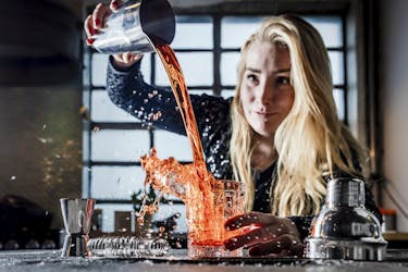 Eine junge Frau vor einer Barkulisse, die eine rote Flüssigkeit aus einem Cocktailshaker in ein Glas schüttet, sodass es spritzt