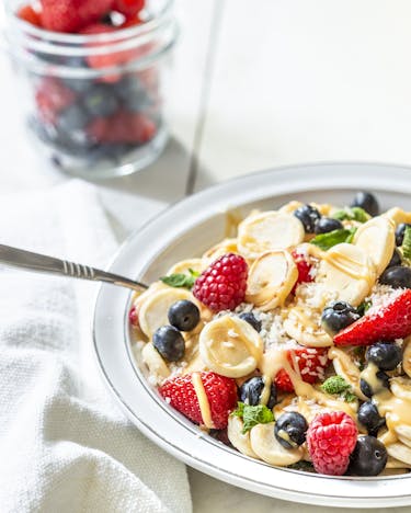 Pancake Cereals – Mini-Pfannkuchen in einer Frühstücksschüssel mit roten Beeren, Mandelmus und Minze, fotografiert seitlich auf hellem Untergrund.
