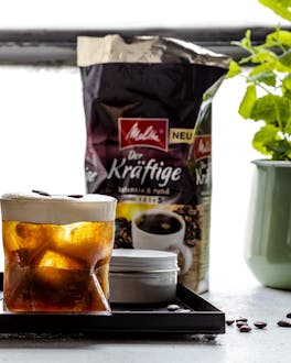 Drei alkoholische Kaffee-Drinks in Tumbler auf Febsterbank mit angestellter Kaffeepackung