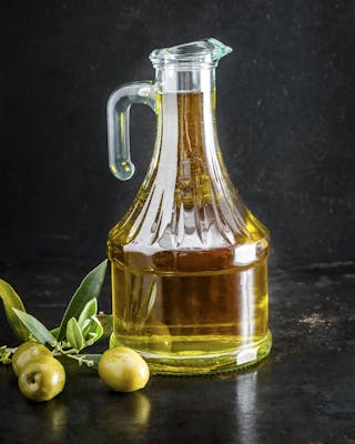 Eine Karaffe mit grünlichem Olivenöl neben frischen grünen Oliven am Zweig vor dunklem Hintergrund