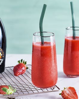 Zwei Cocktailgläser mit eisgekühltem Erdbeer Prosecco Slush auf einem hellen Untergrund neben frischen Erdbeeren und Vanilleschoten vor türkisem Hintergrund