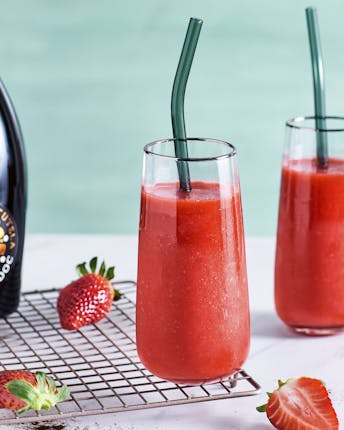 Zwei Cocktailgläser mit eisgekühltem Erdbeer Prosecco Slush auf einem hellen Untergrund neben frischen Erdbeeren und Vanilleschoten vor türkisem Hintergrund
