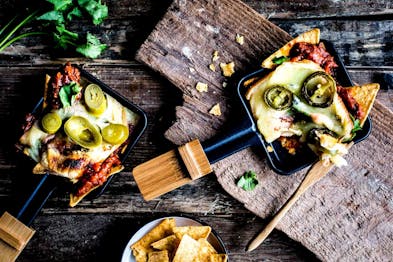 In zwei Raclettepfännchen sind mit Tomatensauce und Käse überbacken, darauf Jalapeñoringe. Die Pfännchen stehen auf dunklem Holzuntergrund.
