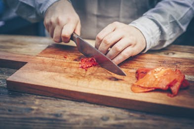 Auf einem Holzbrett wird eine gegrillte Paprika mit einem Messer zerkleinert