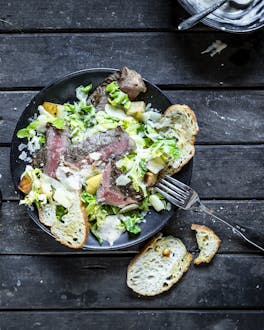 Caesar Salad mit Steakstreifen und Apfel, fotografiert in dunklem Teller auf dunklem Untergrund. Eine Gabel hängt halb im Gericht, ein Crostini liegt daneben, etwas Käse und Salz ist ebenfalls zu sehen.