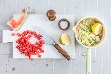 Gewürfelte Wassermelone auf einem weißen Brett. Daneben eine halbe Zitrone, ein großes Küchenmesser und Salz und Pfeffer. Rechts im Bild eine weiße Schale mit Spargelfond