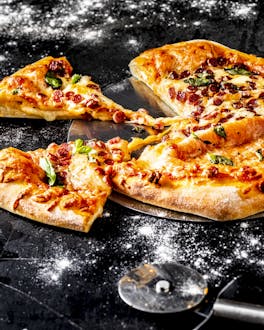 Eine angeschnittene Pinsa, die mit Salat und viel Käse belegt ist, liegt auf einem Pizzaschieber mit Holzgriff vor einem bemehlten dunklen Untergrund neben einem Pizzarad