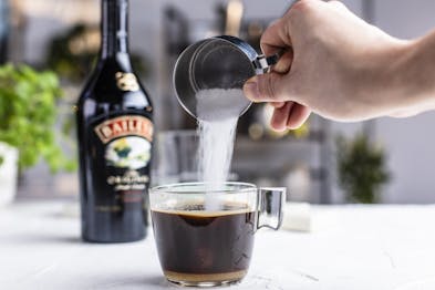 Zucker wird in eine Glastasse mit Espresso geschüttet. Baileys Flasche im Hintergrund.