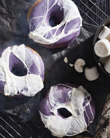 Vier violett glasierte Donuts mit Marshmallow-Spinnennetz neben einer Schale mit weißen Marshmallows auf dunklem Untergrund