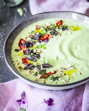 Hellgrüne Suppe in tiefem Teller mit Deko und angelegten Brotscheiben auf lilafarbener Tischdecke