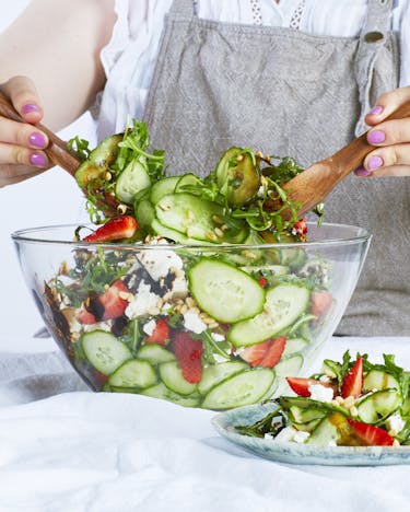In einer Glasschüssel befindet sich Gurkensalat mit Erdbeeren und Feta. Eine Person mischt den Salat mit Besteck
