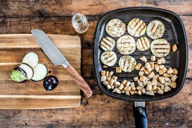 Auberginenscheiben und Putenbrustwürfel werden in einer Grillpfanne angebraten, daneben ein Holzbrett mit großem Messer und geschnittener Aubergine