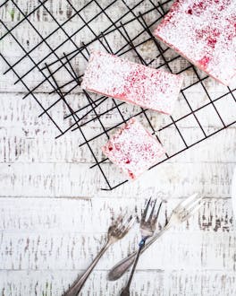 Weiß-pinke Rhabarberschnitten liegen portioniert auf einem weißen Teller. Daneben ist auf einem Gitter die übrige Tarte zu sehen.