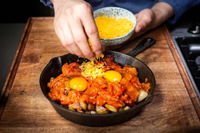 Patats bravas in einer schwarzen Pfanne mit Tomaten und Ei wird mit Käse bestreut