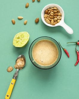 Erdnuss Limetten Dressing Mit Chili in einer Glasschüssel auf grünem Untergrund, daneben eine Schüssel mit Erdnüssen und eine ausgepresste Limette.