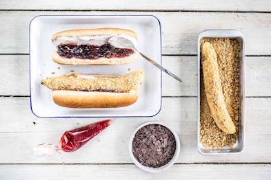 Auf einem Teller liegt ein aufgeschnittenes Hotdog-Brötchen, daneben weitere Zutaten