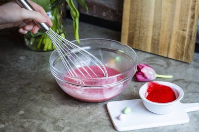 In einer Glasschüssel werden Sahne, weiße Kuvertüre und rote Lebensmittelfarbe zu einer pinken Ganache für einen Drip Cake angerührt.