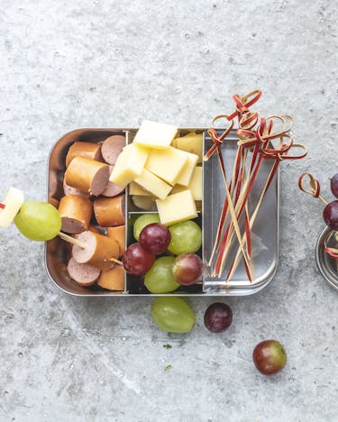 Spieße mit Weintrauben, Wurst und Käse in Metall-Brotbox auf grauem Untergrund