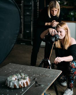 2 Frauen stehen hinter einer Kamera und fotografieren einen Kuchen.