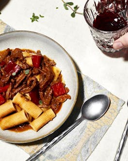 Gulasch mit Pasta auf einem Teller. Daneben liegt Besteck. Eine Hand hält ein Glas mit Wein.