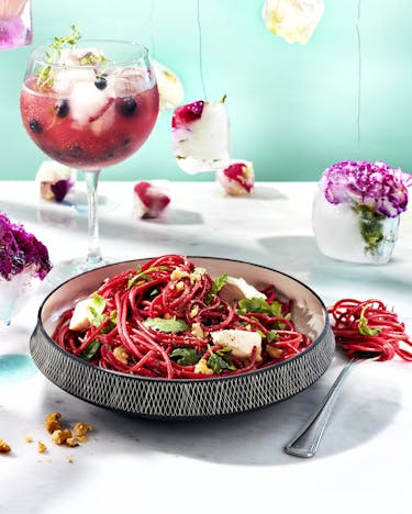 Die tiefroten Spaghetti sind mit Ziegenkäse und Rucola in einer Schüssel vor einem roten Longdrink, mit Blüten gefüllten Eiswürfeln angerichtet